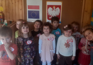Dzieci stoją przy tablicy z symbolami narodowymi, mają przypięte kotyliony.